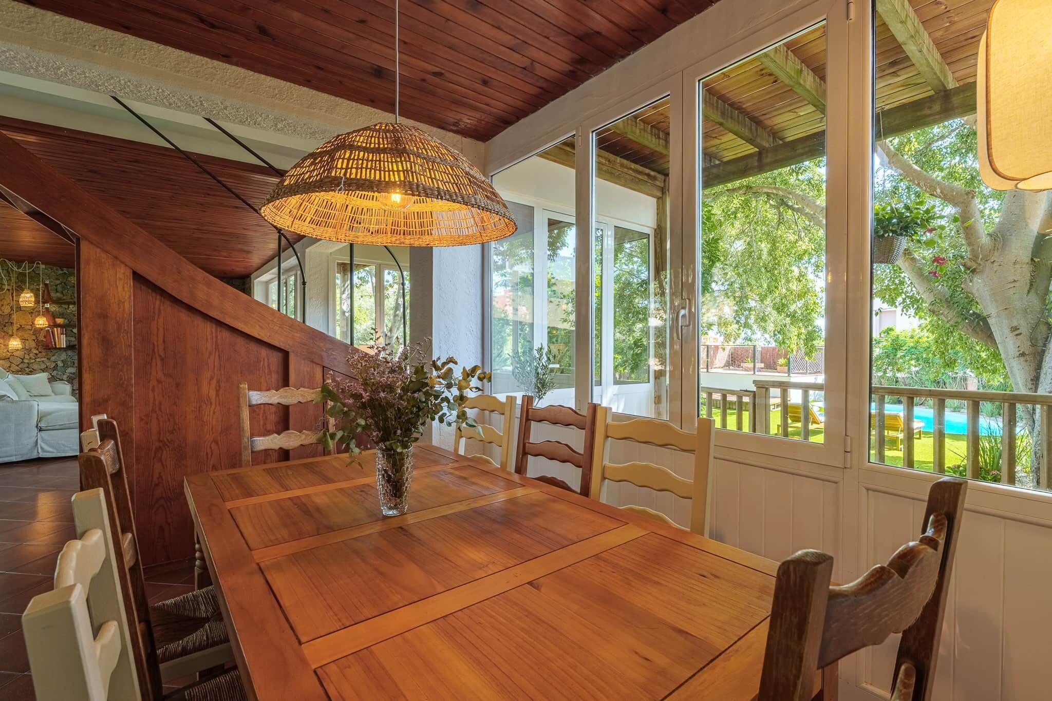 A dining area with a wooden table and chairs, a hanging light fixture, and a view of the outdoors.