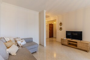 A living room with a grey sofa, a flat-screen TV on a wooden stand, and a hallway leading to a door. - #413 Charming 1-Bedroom Apartment | Salgados, Albufeira (Albufeira)