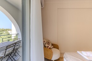 Un balcón con una mesa y sillas, una silla de mimbre con cojín, y una cama con una toalla blanca.