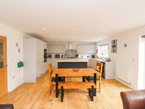 A modern kitchen with white cabinets, a central island, and a dining area with a wooden table and chairs. - 95 Cae America (Llanfairfechan)