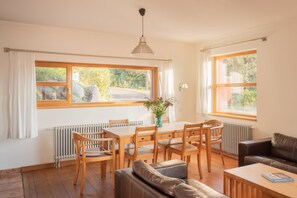 Et værelse med spisebord, stole, sofa, sofabord og et vindue med udsigt til grøn vegetation.