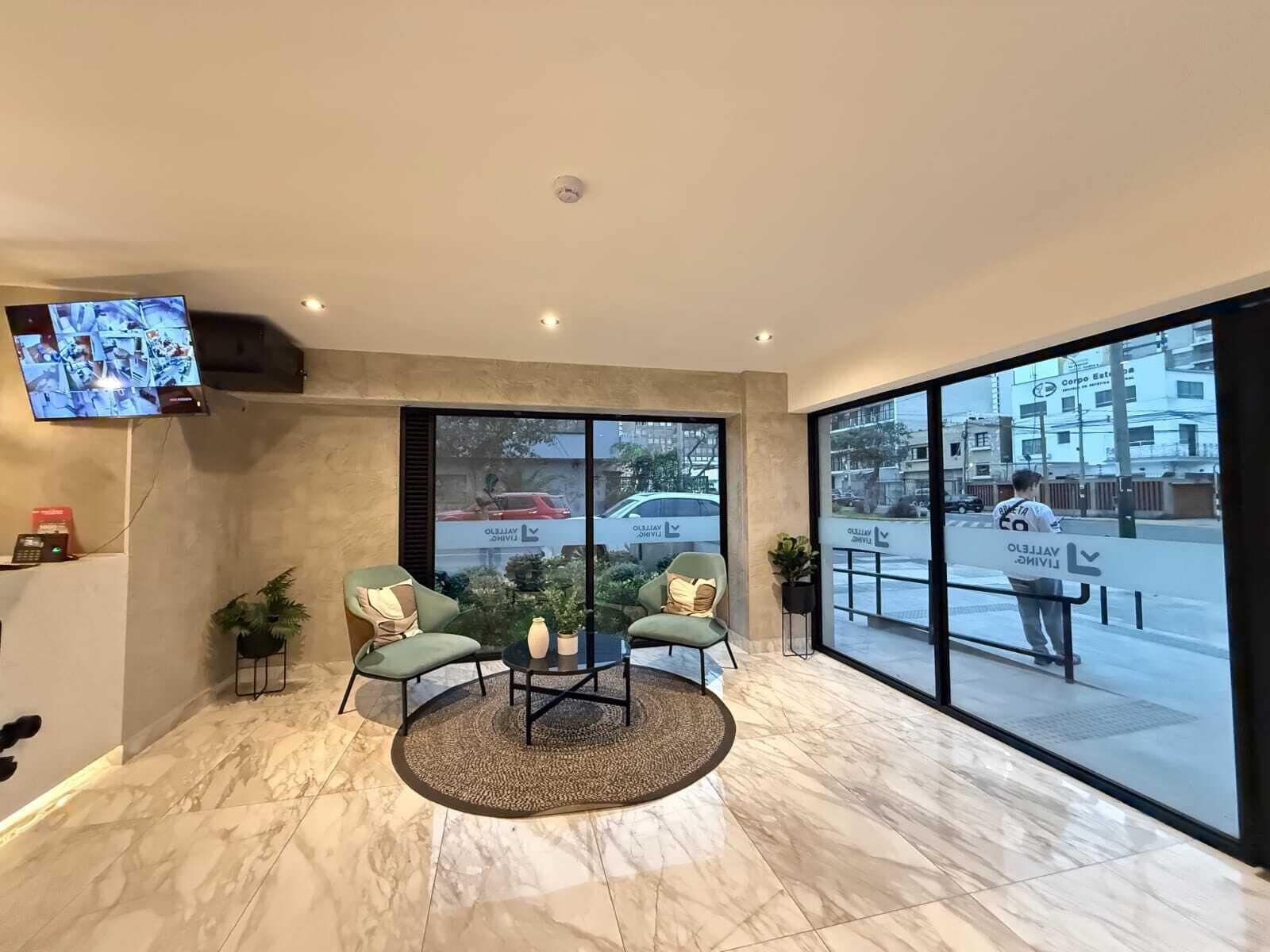 Un vestíbulo moderno con piso de mármol, dos sillones verdes, una mesita y un amplio ventanal con vistas a la calle.