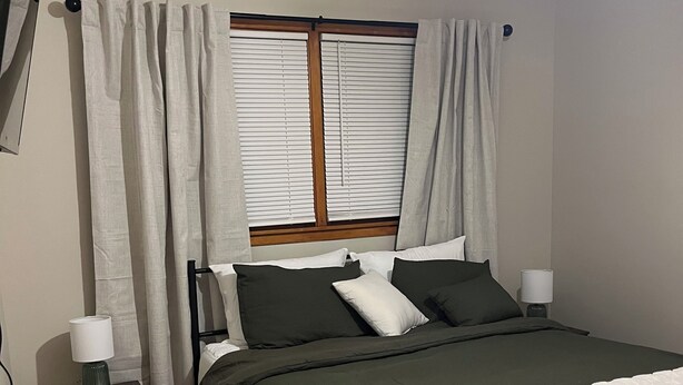 Un dormitorio con cama, mesita de noche con lámpara, ventana con persianas y ventilador de techo.