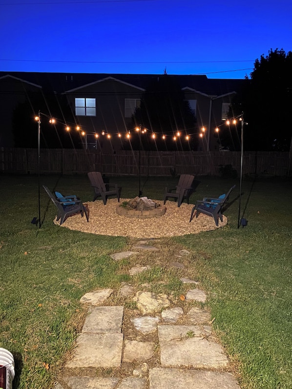 Un patio trasero con una fogata, asientos exteriores y luces colgantes.
