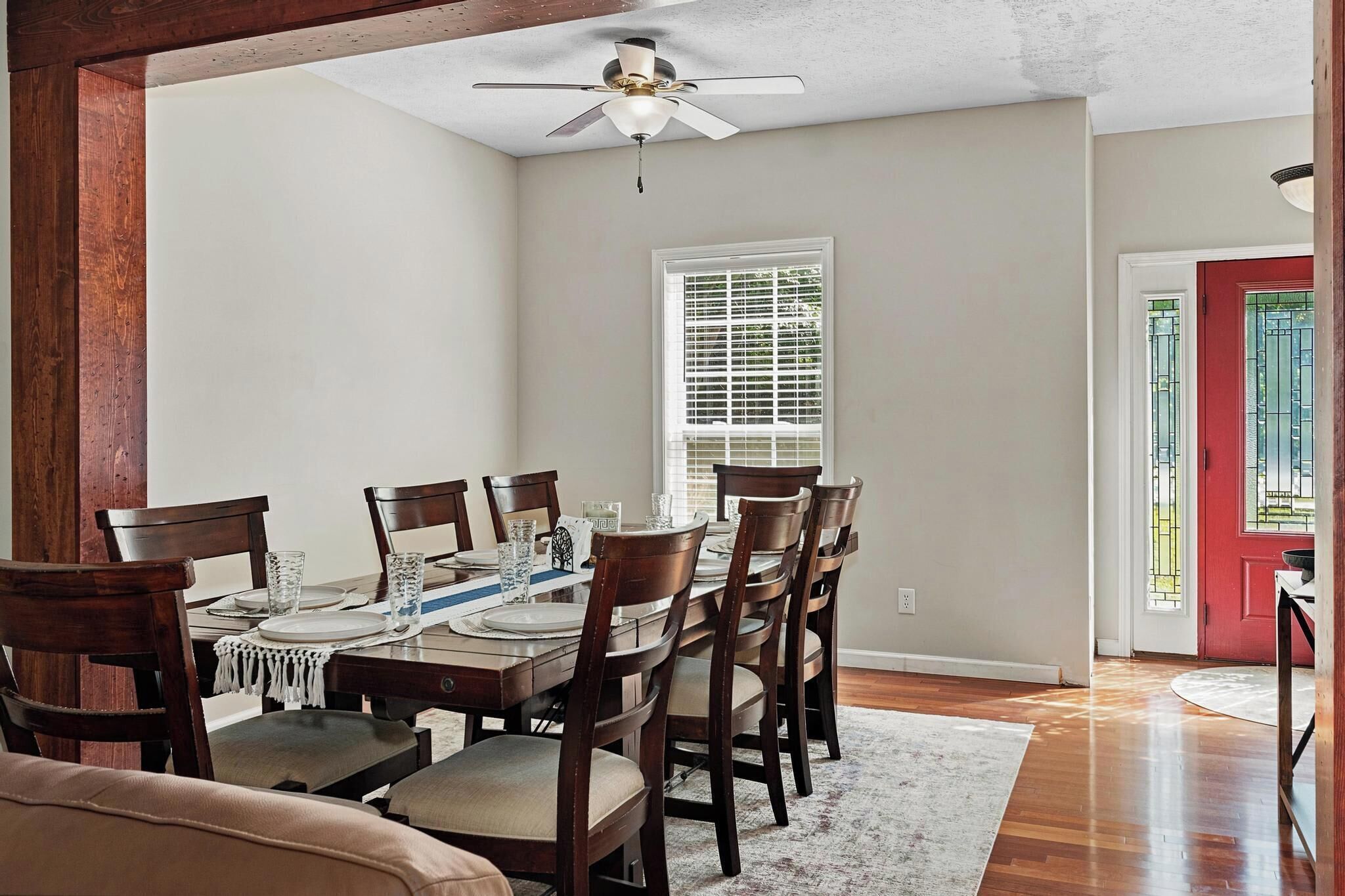 A dining area with a table set for six, wooden chairs, a ceiling fan, a window with blinds, and a red door.