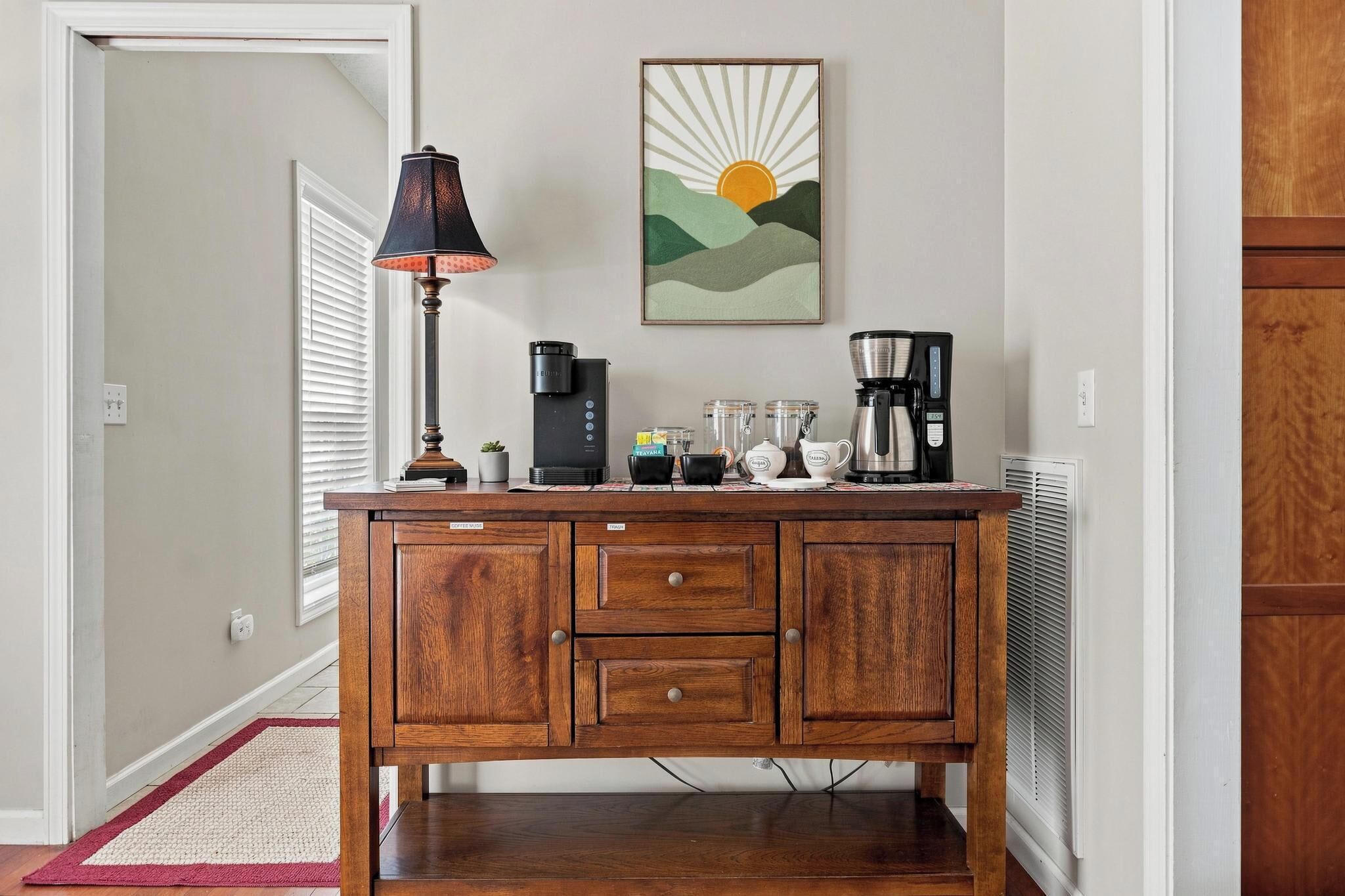 A wooden coffee station with a lamp, coffee maker, and storage drawers.