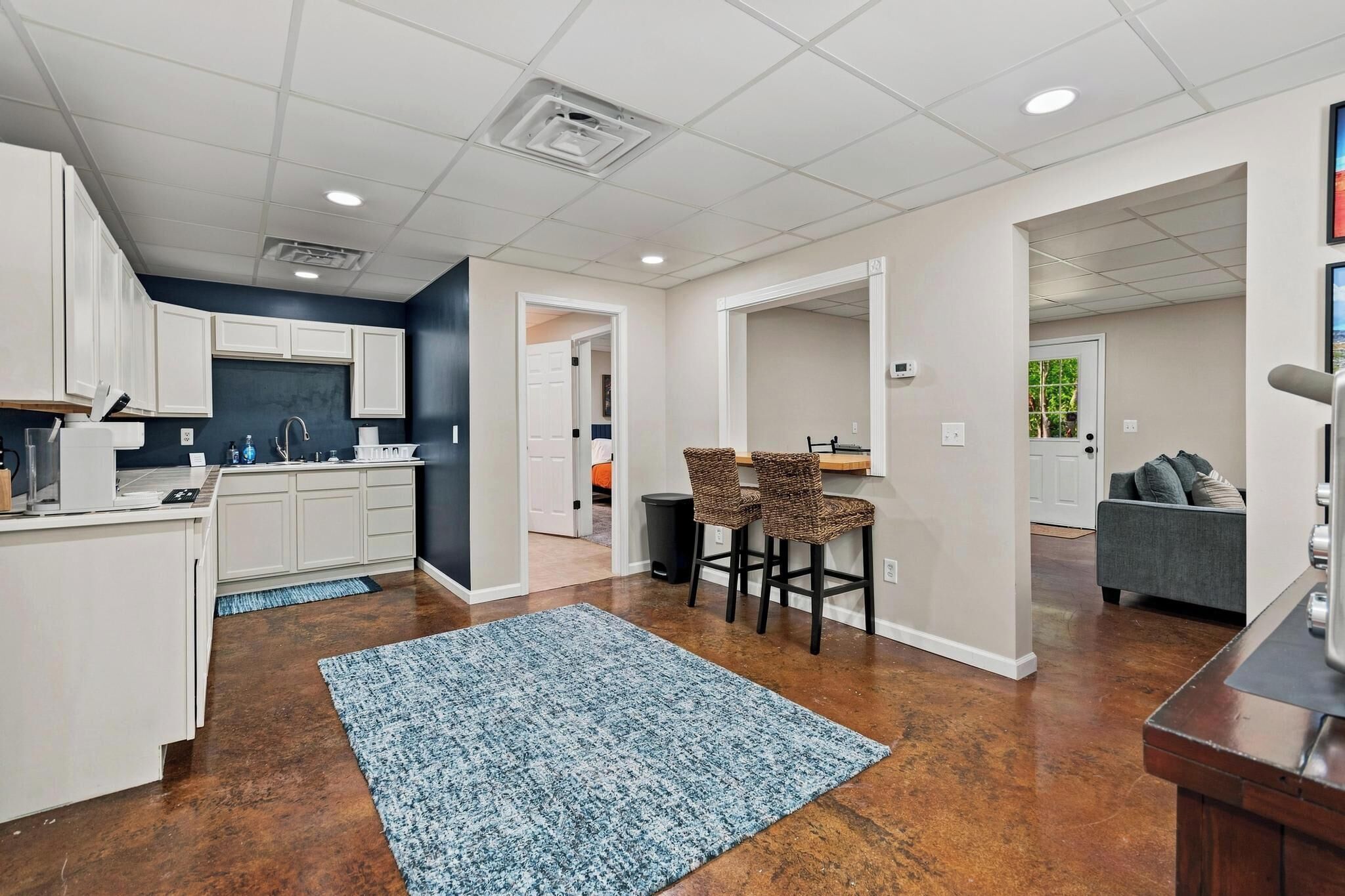 A modern kitchen with white cabinets, a dining area with a blue rug, and a living room with a gray sofa.