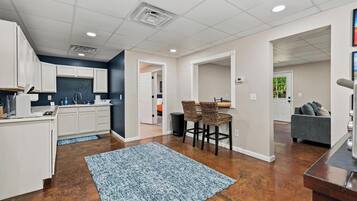A modern kitchen with white cabinets, a dining area with a blue rug, and a living room with a gray sofa.
