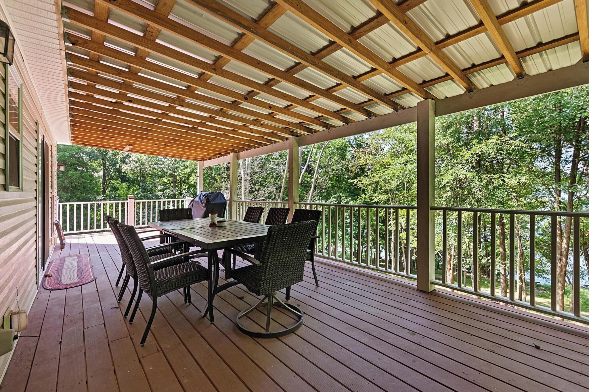 A covered outdoor patio with a table and chairs, surrounded by a wooden deck and a railing.