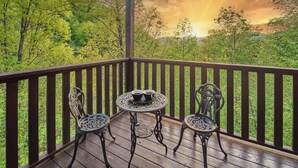 Um deck de madeira com duas cadeiras de metal decoradas e uma mesa redonda pequena, com um fundo de vegetação exuberante e um pôr do sol.