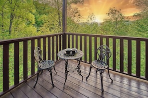 A wooden deck with two ornate metal chairs and a small round table, set against a backdrop of lush greenery and a sunset. - Peaceful Cabin with Hot Tub, Game Room & Mtn Views (Sevierville)