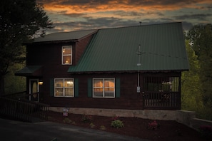 A two-story wooden house with a green metal roof, a front porch, and a small garden. - Peaceful Cabin with Hot Tub, Game Room & Mtn Views (Sevierville)
