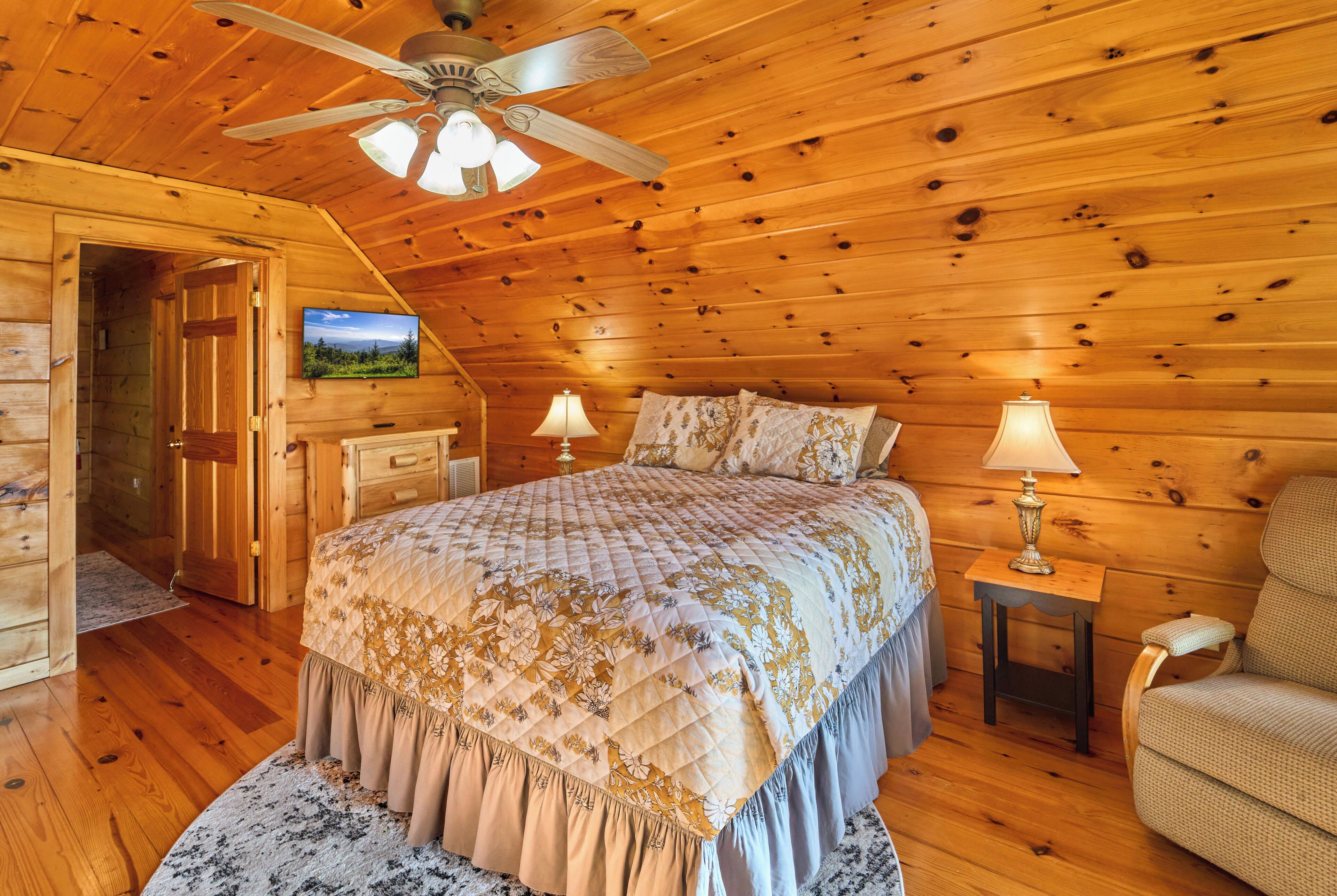 Une chambre à coucher aménagée dans un chalet en rondins, avec un lit, deux lampes de chevet, un ventilateur de plafond et un téléviseur fixé au mur.