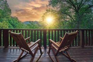 A wooden deck with two rocking chairs facing a scenic view of a forest and mountains during sunset. - Peaceful Cabin with Hot Tub, Game Room & Mtn Views (Sevierville)