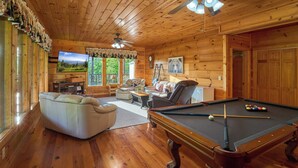 Interior de cabana de madeira com mesa de bilhar, sofá, televisão e lareira.
