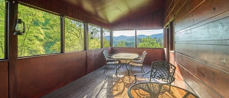 Terraza de madera con mesa y sillas, ventanas amplias y vista a la vegetación.