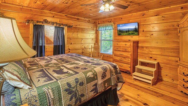 Quarto de cabana de madeira com cama, ventilador de teto, janela com cortinas, cômoda e um banquinho pequeno.
