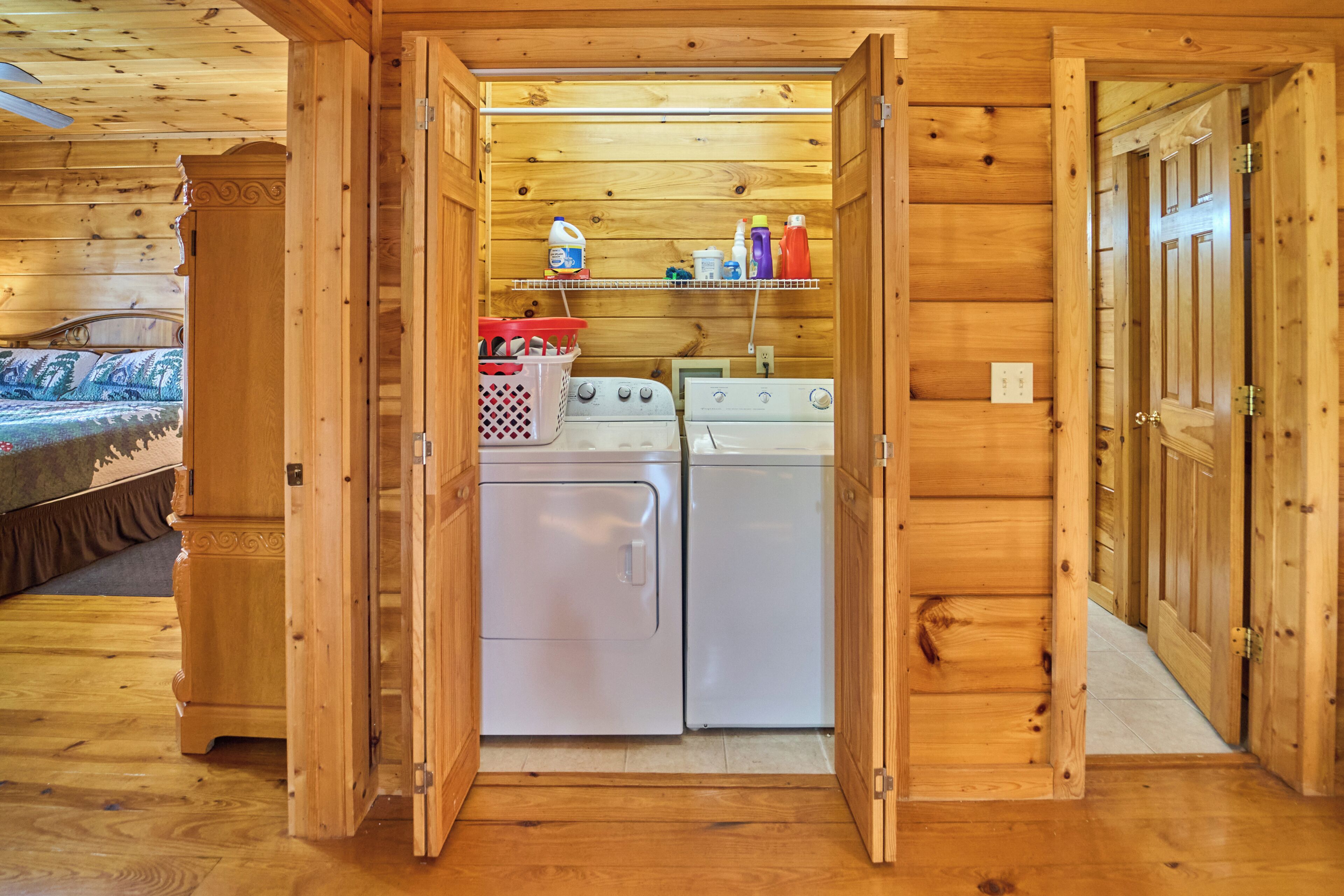 A compact laundry area with two washing machines, a shelf with cleaning supplies, and a bed with a patterned bedspread.