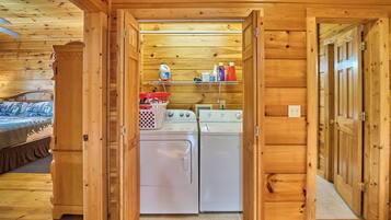 A compact laundry area with two washing machines, a shelf with cleaning supplies, and a bed with a patterned bedspread.