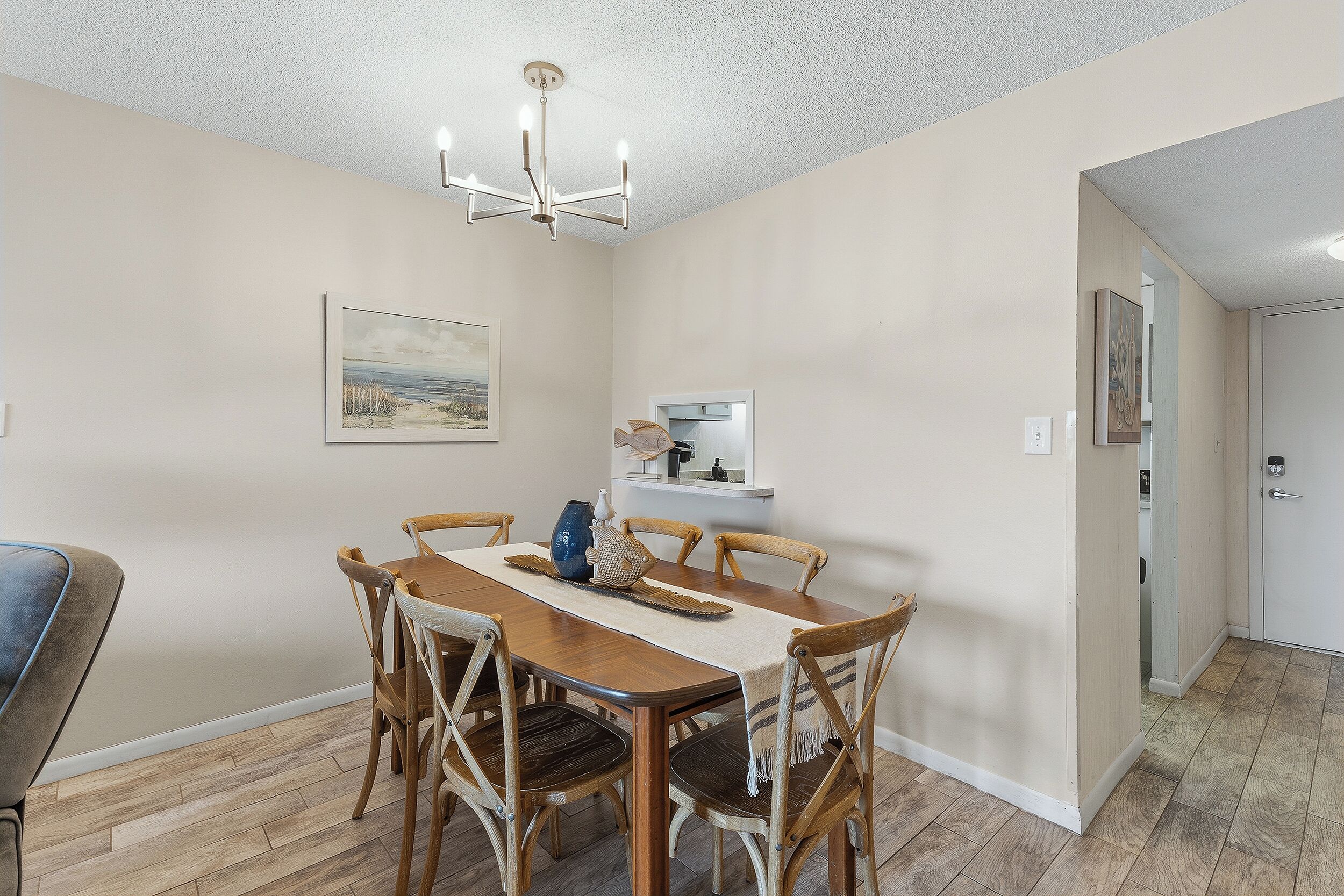 Un coin repas comprenant une table en bois, six chaises, un lustre, un tableau accroché au mur et une petite étagère avec quelques objets.