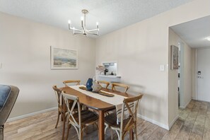 A dining area with a wooden table, six chairs, a chandelier, a painting on the wall, and a small shelf with items. - Ocean Views & Coastal Comfort | Ocean Breeze Hideaway | Sea Coast Gardens II 511 (New Smyrna Beach)