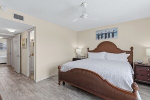 A bedroom with a wooden bed, a nightstand, a wall-mounted lamp, and a ceiling fan. - Ocean Views & Coastal Comfort | Ocean Breeze Hideaway | Sea Coast Gardens II 511 (New Smyrna Beach)