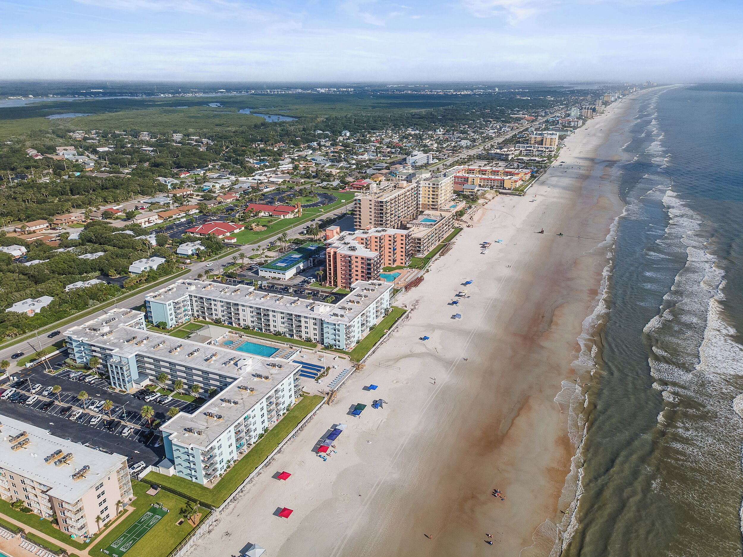 An aerial view of a coastal city with a beachfront hotel, including a swimming pool and beach umbrellas.