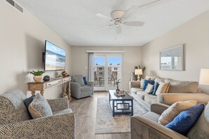 A living room with a ceiling fan, a television, a sofa, a chair, and a coffee table. - Ocean Views & Coastal Comfort | Ocean Breeze Hideaway | Sea Coast Gardens II 511 (New Smyrna Beach)