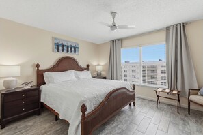 Ein Hotelzimmer mit einem großen Bett, einem Nachttisch, einer Lampe, einem Fenster mit Vorhängen und einem Deckenventilator.