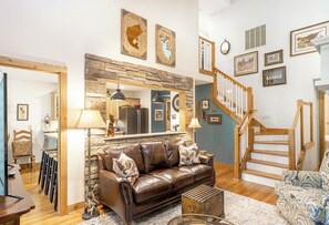 A living room with a stone wall, a brown leather sofa, a wooden staircase, and a kitchen in the background. - Windchime 7A- Mtn Townhome, Indoor/Outdoor Pool & Hot Tub (Boone)