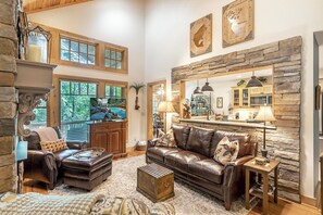 A spacious living room with a stone fireplace, leather sofa, and a large window overlooking greenery. - Windchime 7A- Mtn Townhome, Indoor/Outdoor Pool & Hot Tub (Boone)