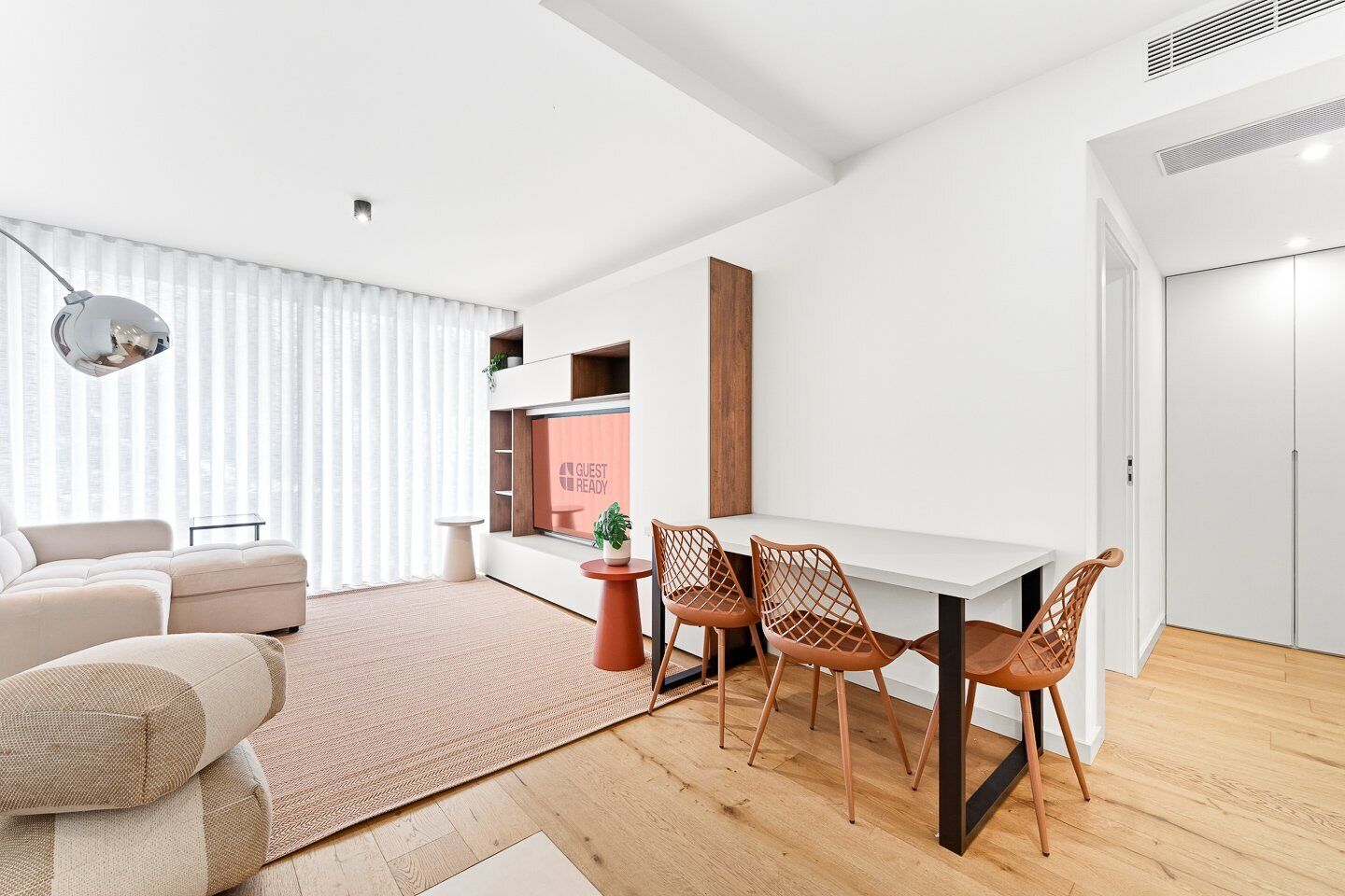 Ein modernes Wohnzimmer mit einer Couch, einem Esstisch mit Stühlen, einem kleinen Beistelltisch mit Pflanze und einem Fenster mit Jalousien.