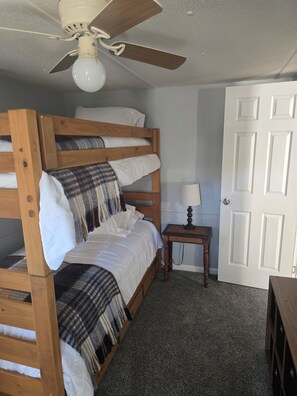 Una cama individual superpuesta con ventilador de techo, una mesita de noche con lámpara, y una puerta en una habitación con piso alfombrado.