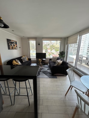 Un salon moderne avec un canapé, une table à manger et un téléviseur. De grandes fenêtres offrent une vue sur la ville.
