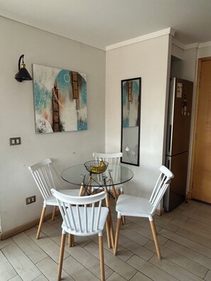 Een eetgedeelte met een glazen tafel, omringd door witte stoelen, een wandlamp en twee abstracte schilderijen.