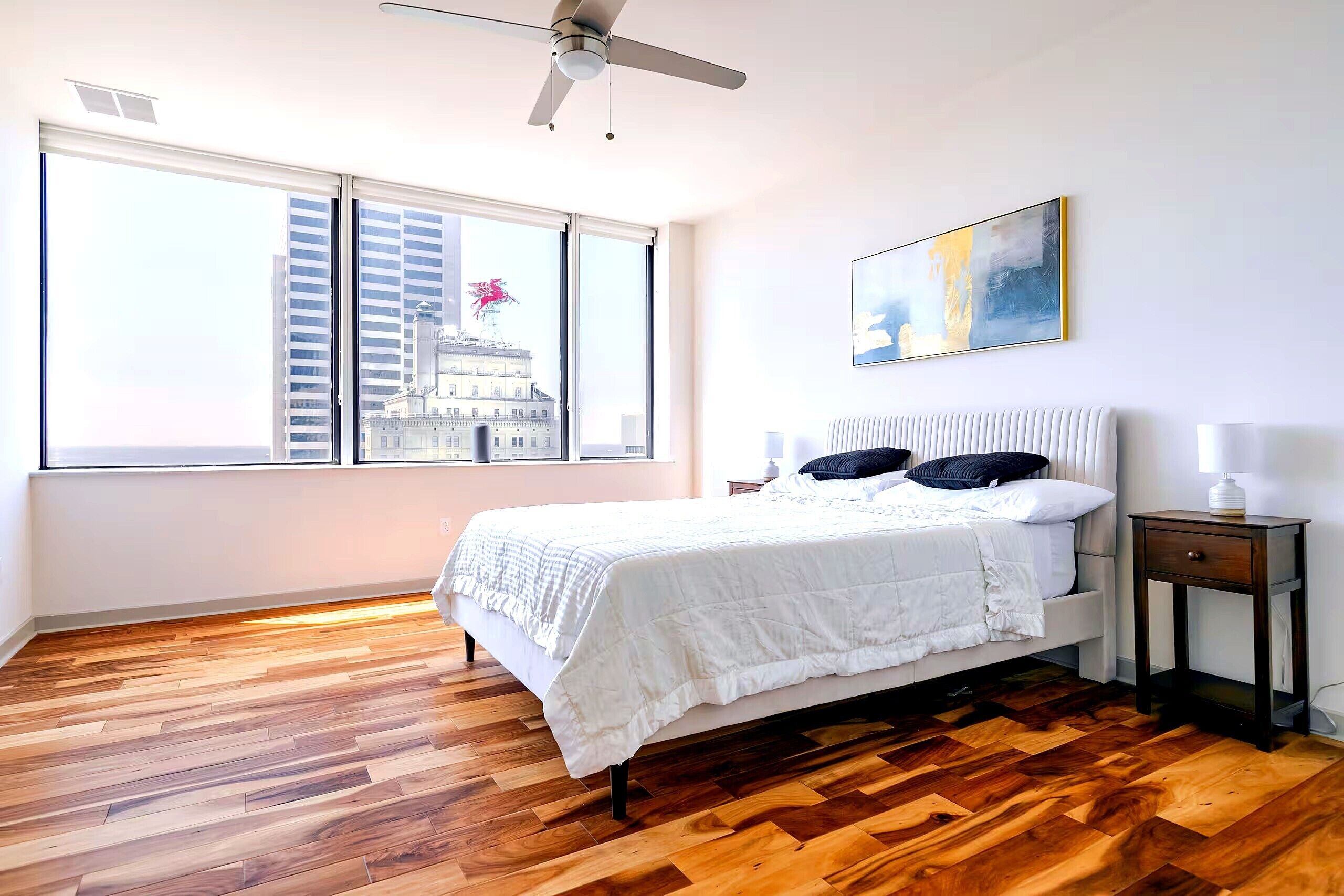 A bedroom with a large window, a bed, a bedside table, and a ceiling fan.
