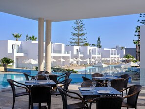 A poolside dining area with tables and chairs, a pool, and modern buildings in the background. - Panthea Resort (Ayia Napa)