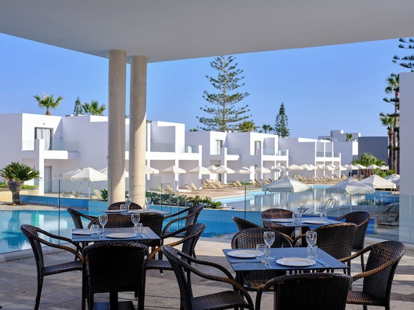 Un área de comedor junto a la piscina con mesas y sillas, una piscina y edificios modernos al fondo.