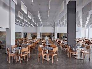 Un comedor amplio con varias mesas y sillas, una zona de cocina central y ventanas grandes que dejan entrar luz natural.