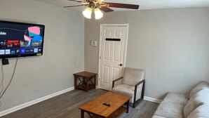 A living room with a television mounted on the wall, a ceiling fan, a wooden coffee table, a beige armchair, and a sofa. - Roomy 5BR Near I-95 & Fort Bragg - Family, Group & Work Stays (Fayetteville)