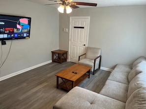 Een woonkamer met een aan de muur bevestigde televisie, een plafondventilator, een houten salontafel, een beige fauteuil en een bank.