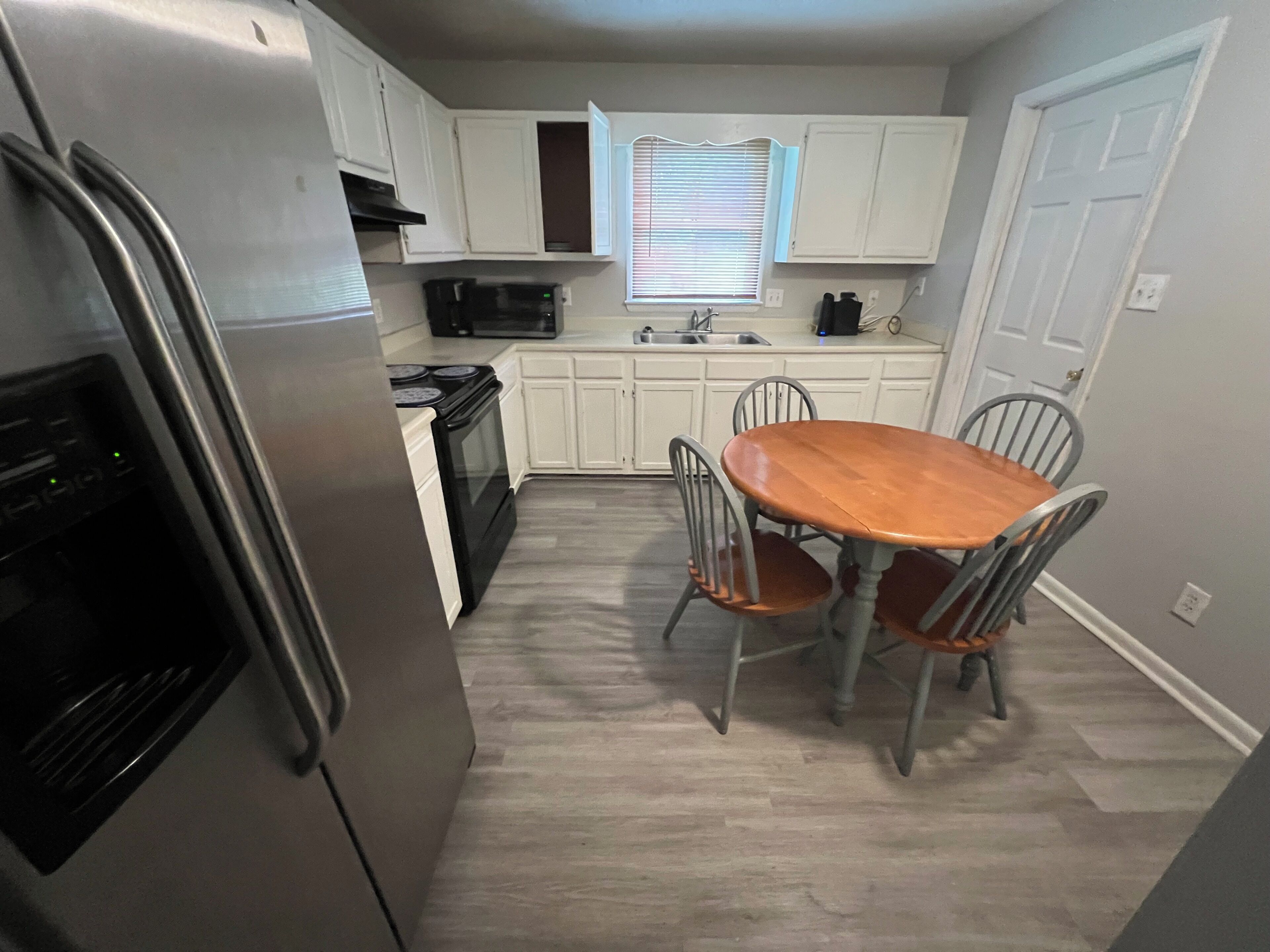 A compact kitchen with white cabinets, a stainless steel refrigerator, a microwave, an oven, a sink, and a dining table with four chairs.