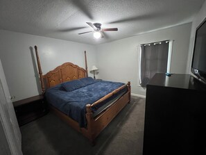 Een slaapkamer met een houten bed, een plafondventilator, een raam met gordijnen en een televisie op een dressoir.
