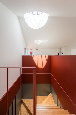 Un escalier avec des marches en bois et des rampes rouges, menant à un palier avec un mur blanc et un luminaire au plafond.