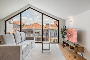A modern living room with a large window, a grey sofa, a small table, and a TV on a wooden stand. - GuestReady - Joy Apartments - F (Porto)