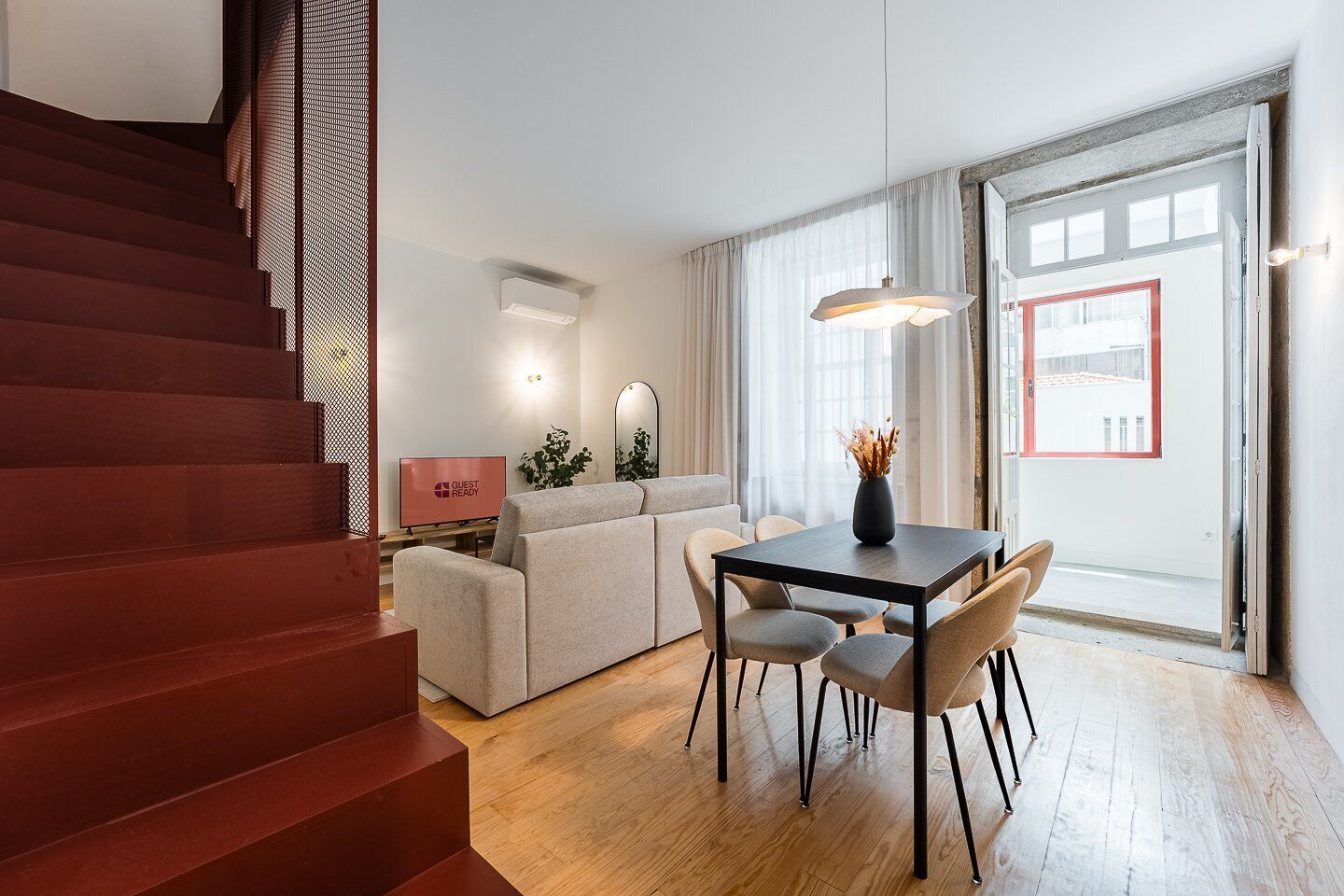 A modern living room with a sofa, dining table, and chairs. There is a staircase on the left and a large window on the right.