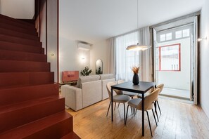 Ein modernes Wohnzimmer mit einer Couch, einem Esstisch und Stühlen. Eine Treppe befindet sich links und ein großes Fenster rechts.