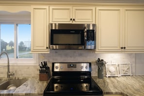 A kitchen with white cabinets, a stainless steel microwave, a black stove, and a window with a view of the outdoors. - Just Beachy, Private Pool, Beach Front (Saint George Island)