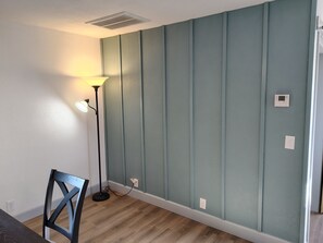 A room with a wooden floor, a standing lamp, a chair, and a wall with a built-in thermostat.