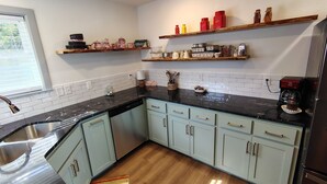A kitchen with light blue cabinets, black countertops, and a wooden floor. There are two shelves above the counter with various kitchen items.